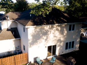 Two-story home with light-colored siding and dark roof, backyard patio, and trees casting shadows—illustrating residential siding installation context.